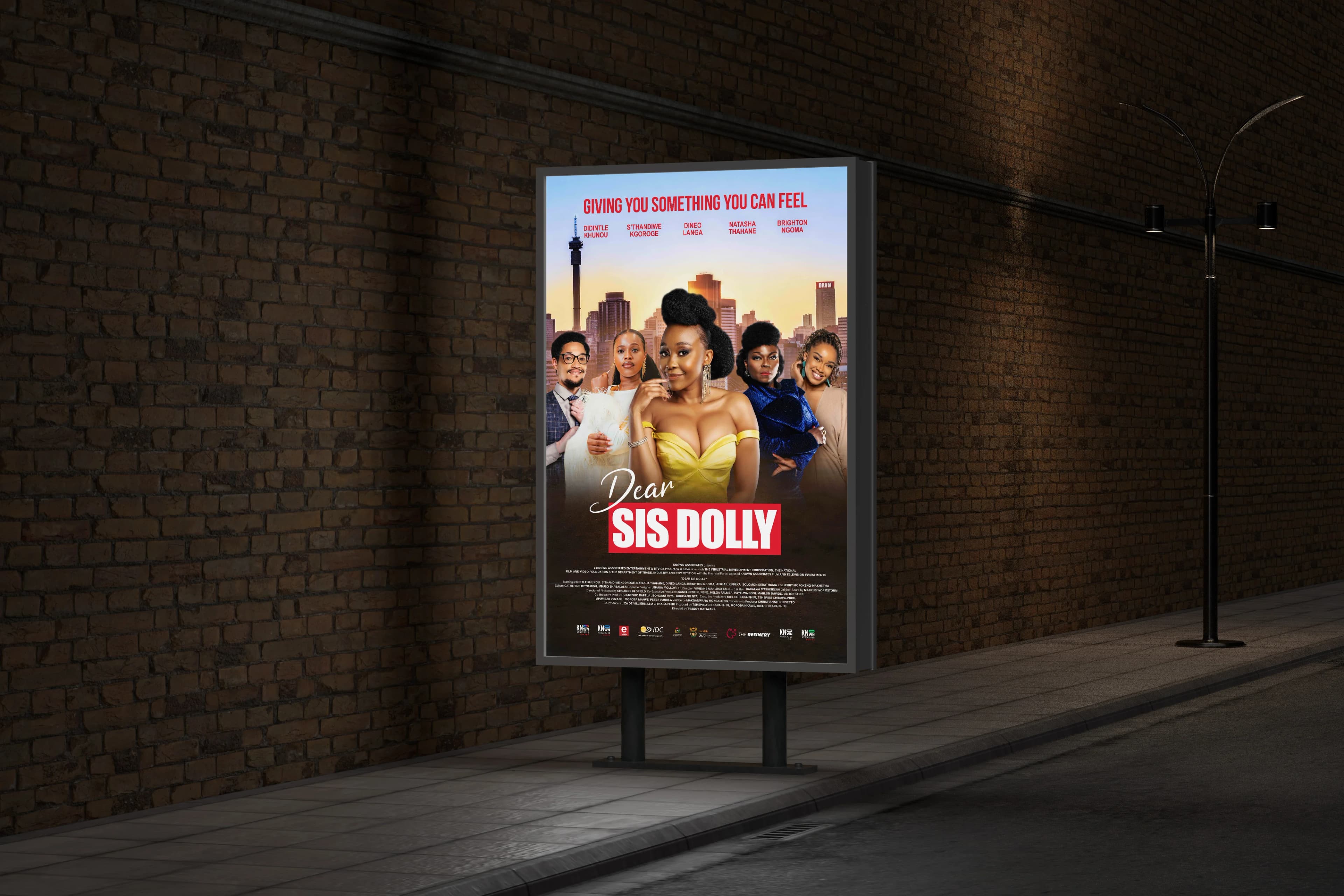 Portrait Dear Sis Dolly one-sheet presented in an illuminated street-side poster case beside a brick wall at night, with the full credit block along the base.